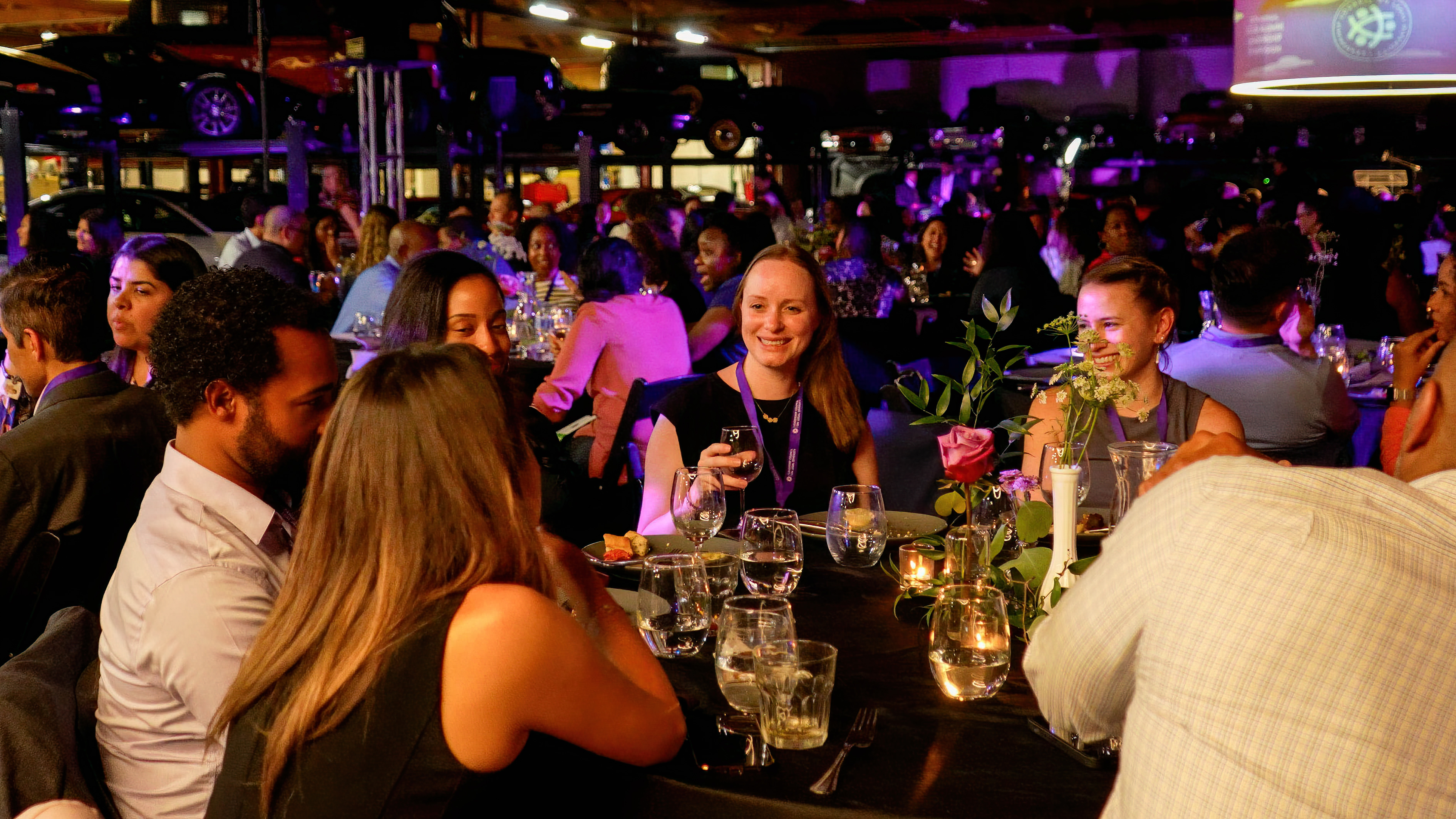 Corporate gala networking reception — professionals in blazers gathered at bar with purple uplighting and classic cars
